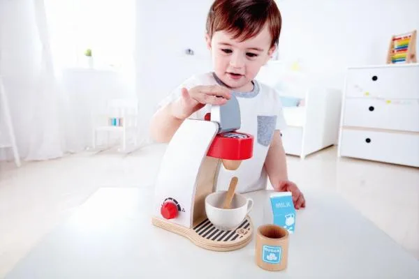 Kind spielt mit Holz-Kaffeemaschine, legt Pad ein und hat eine Tasse mit Holzlöfel, Milchtüte und Zuckerdose griffbereit.