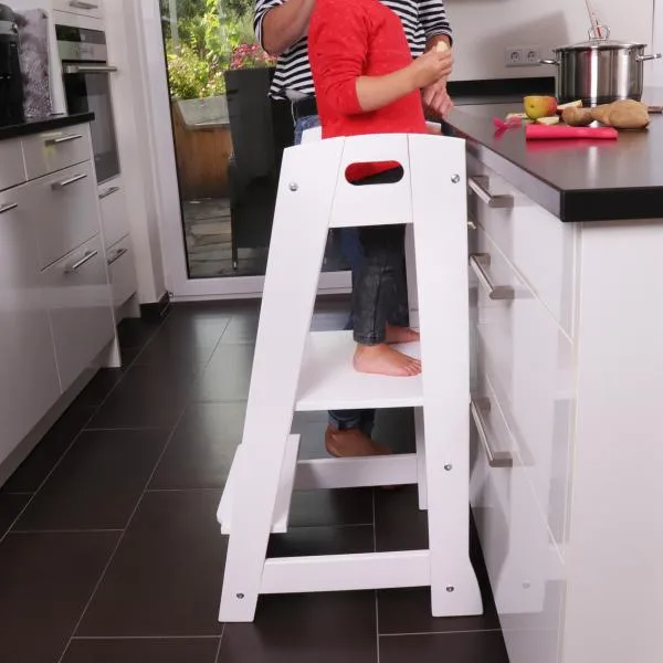 Kleinkind steht auf einem weißen, stabilen Lernturm an der Küchenzeile und schaut der Mutter beim kochen zu.